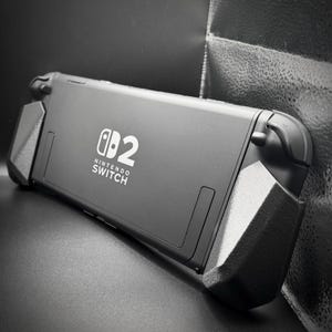 Puede incluir: Consola Nintendo Switch negra con mandos acoplados. La consola tiene el logotipo de Nintendo Switch y el número "2" en blanco. Los mandos son negros y tienen un agarre texturizado.