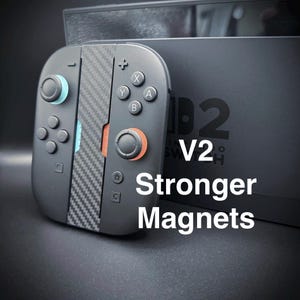 May include: Black game controller with carbon fibre accents and text that reads "V2 Stronger Magnets". The controller has blue and orange accents on the joysticks and buttons. The background is a black box.
