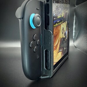 Puede incluir: Mando de juegos negro con un joystick azul y una funda negra que sujeta una tableta. La tableta muestra una imagen colorida. El mando tiene varios botones y una cruceta. La funda tiene un bot&oacute;n de encendido y controles de volumen.