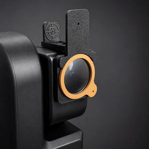 Puede incluir: Un dispositivo negro con una lente circular y un marco naranja. Una pequeña mira telescópica está montada encima de la lente. El dispositivo está conectado a un objeto negro más grande, posiblemente una cámara o una herramienta similar.