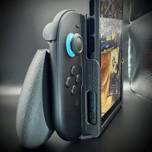 May include: A black gaming controller with a blue accent light, attached to a tablet or phone. The device is held in a textured gray holder, designed for handheld gaming. The screen displays a colorful image.