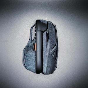 Puede incluir: Un ratón para juegos negro y gris con un controlador adjunto. El ratón tiene una superficie texturizada y un diseño ergonómico. El controlador tiene un botón naranja y un joystick negro. El fondo es gris liso.