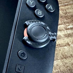Könnte beinhalten: Nahaufnahme eines schwarzen Handheld-Spielgeräts mit einem schwarzen Joystick und Tasten mit den Bezeichnungen Y, B, A. Der Joystick hat eine schwarze kreisförmige Oberseite mit roter Mitte. Das Gerät hat eine Home-Taste und eine Zurück-Taste.