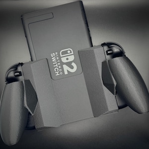 Puede incluir: Una consola Nintendo Switch negra est&aacute; insertada en un agarre de controlador gris. El logotipo de Nintendo Switch es visible en la consola. El agarre del controlador tiene dos asas a cada lado.