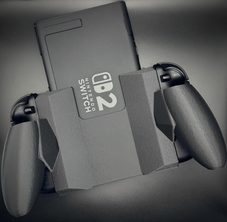 Puede incluir: Una consola Nintendo Switch negra est&aacute; insertada en un agarre de controlador gris. El logotipo de Nintendo Switch es visible en la consola. El agarre del controlador tiene dos asas a cada lado.