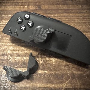 Peut inclure: Manette de jeu noire avec des boutons blancs marqués A, B, X et Y. La manette a un motif en fibre de carbone et un composant articulé séparé. L'appareil repose sur une surface en bois.