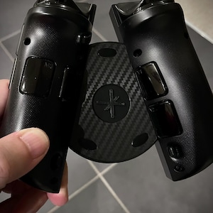 May include: Black gaming controller with a carbon fiber center piece. The controller has a textured grip and several buttons. The center piece has a cross-shaped design. The controller is held in a person's hand.