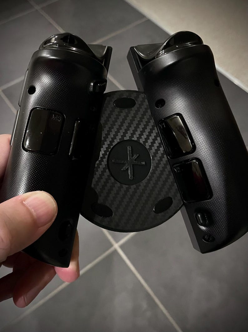 May include: Black gaming controller with a carbon fiber center piece. The controller has a textured grip and several buttons. The center piece has a cross-shaped design. The controller is held in a person's hand.