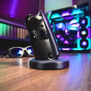 May include: A black gaming controller resting on a dark blue stand. The controller has two buttons and a textured grip. The background includes a computer with colorful lights and a pair of glasses.