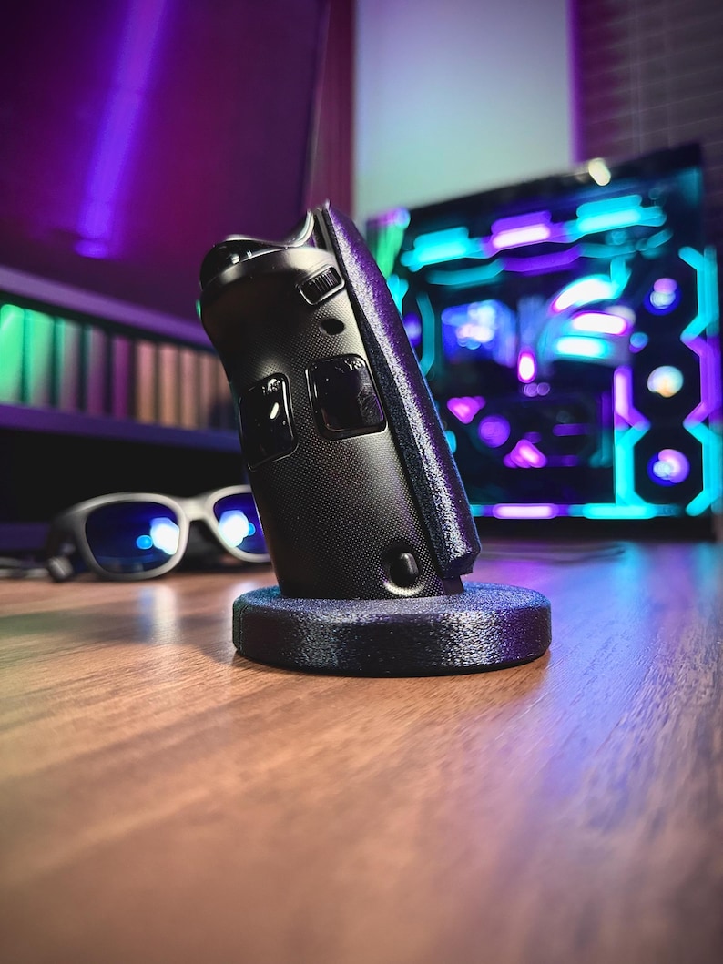May include: A black gaming controller resting on a dark blue stand. The controller has two buttons and a textured grip. The background includes a computer with colorful lights and a pair of glasses.
