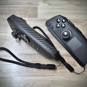 May include: Two black game controllers with a carbon fiber texture and a wrist strap. One controller has a joystick and directional pad, while the other has buttons and a touchpad. The controllers are on a wooden surface.
