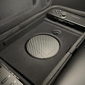 May include: Black carrying case with a zipper, holding a black handheld gaming device and a round carbon fiber cover. The case is open, revealing the interior with a controller and a circular compartment.