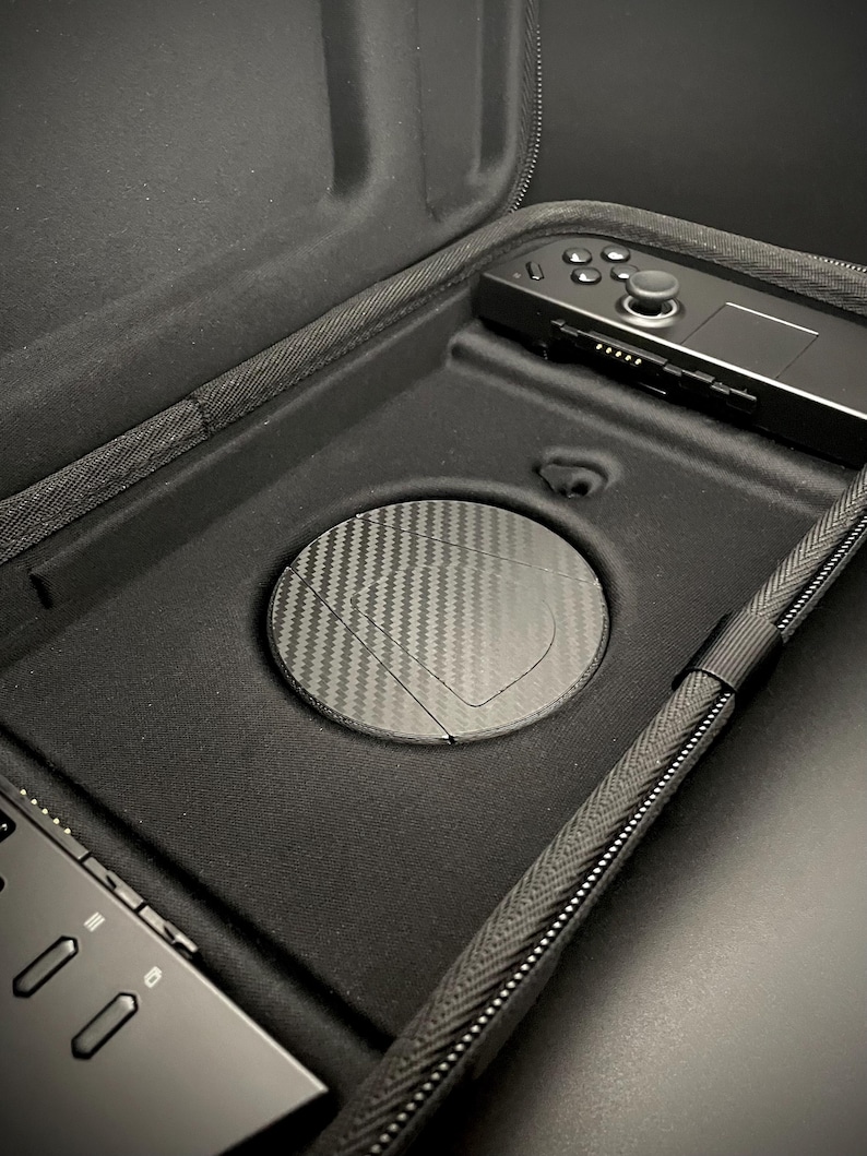 May include: Black carrying case with a zipper, holding a black handheld gaming device and a round carbon fiber cover. The case is open, revealing the interior with a controller and a circular compartment.