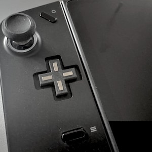 May include: A close-up shot of a black handheld games console. It has a textured analogue stick, a directional pad, and several buttons. The screen is black and reflective. The design is sleek and modern, with a focus on functionality.