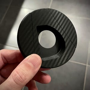 May include: A black, circular object with a carbon fiber weave pattern. The object has a large, D-shaped opening in the center and is held in a person's hand. The object appears to be made of a composite material.