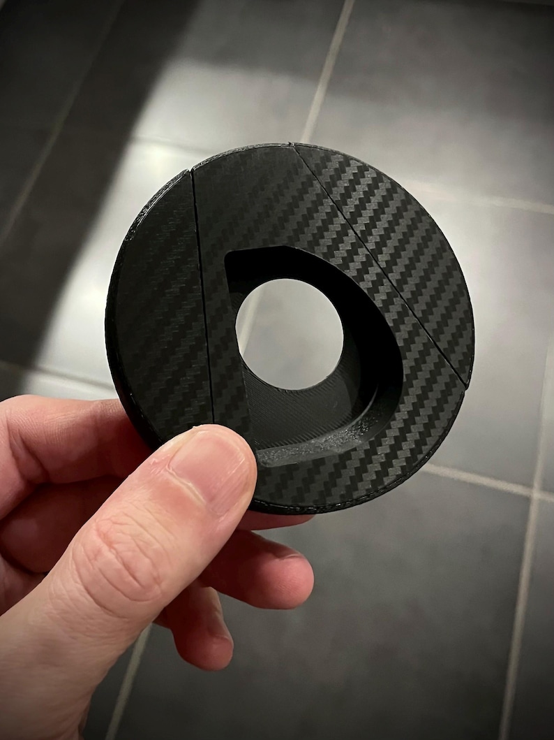 May include: A black, circular object with a carbon fiber weave pattern. The object has a large, D-shaped opening in the center and is held in a person's hand. The object appears to be made of a composite material.