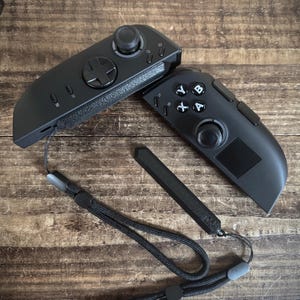 May include: Two black game controllers with buttons, joysticks, and wrist straps on a wooden surface. The controllers have a modern design, with a focus on functionality and ergonomics. The wrist straps are black and attached to the controllers. The wooden surface has a natural grain.