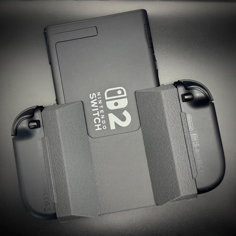 Puede incluir: Una consola Nintendo Switch negra con mandos Joy-Con adjuntos. La consola tiene el logotipo de Nintendo Switch y el n&uacute;mero 2 en blanco. Los mandos Joy-Con est&aacute;n a ambos lados de la consola.
