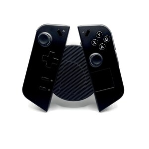 May include: Black gaming controller with a circular button layout and a directional pad. The controller is split into two halves, resting on a textured black circular base. The background is white.