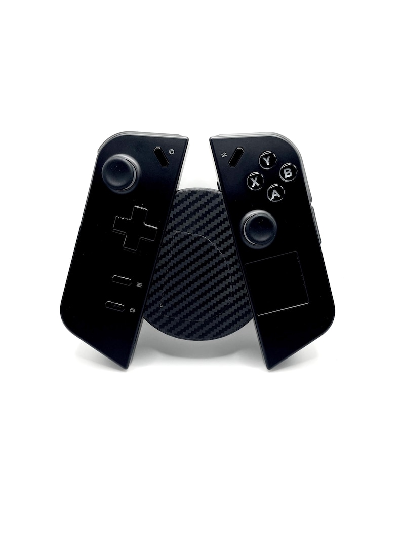 May include: Black gaming controller with a circular button layout and a directional pad. The controller is split into two halves, resting on a textured black circular base. The background is white.