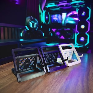 May include: Three phone stands in black, purple, and white. The stands are made of a textured plastic and have a honeycomb design. The stands are angled to hold a phone upright.