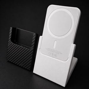 May include: A white wireless charger with a circular charging pad and a stand, alongside a black carbon fibre phone holder. The charger has a minimalist design against a dark background.
