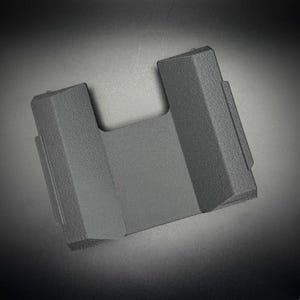 Puede incluir: Un soporte para tel&eacute;fono de pl&aacute;stico gris oscuro texturizado con una abertura en forma de U. El soporte tiene una base rectangular y dos lados verticales en &aacute;ngulo. La superficie tiene una textura ligeramente rugosa y el dise&ntilde;o es minimalista y moderno.