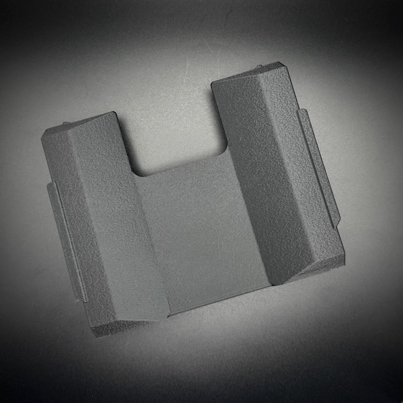 Puede incluir: Un soporte para tel&eacute;fono de pl&aacute;stico gris oscuro texturizado con una abertura en forma de U. El soporte tiene una base rectangular y dos lados verticales en &aacute;ngulo. La superficie tiene una textura ligeramente rugosa y el dise&ntilde;o es minimalista y moderno.