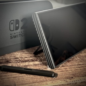 以下が含まれることがあります： カーボンファイバー模様のトリムが付いた黒いNintendo Switchコンソール。コンソールはスタンドで支えられています。黒いスタイラスが前景にあり、Nintendo Switchのロゴがコンソールのベースに表示されています。