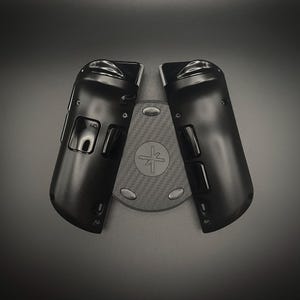 May include: Black gaming controller accessories, featuring a textured surface and a central base with a circular design and a symbol. The controller components are positioned on a dark gray background.