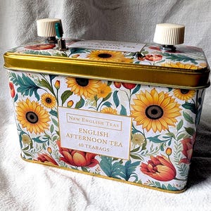 May include: A rectangular metal tea tin featuring a floral pattern of yellow sunflowers and red tulips on a white background. The label reads New English Teas English Afternoon Tea 40 Teabags. Two white plastic knobs are attached to the top lid.