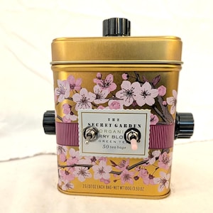 May include: A gold tea tin decorated with pink cherry blossom flowers. The tin has black knobs and a switch. The label reads "The Secret Garden Organic Cherry Blossom Green Tea" with 50 tea bags. The net weight is 100g.