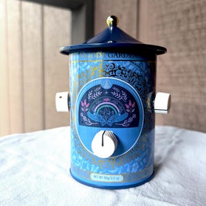 Puede incluir: Una lata cilíndrica de metal azul con una tapa cónica rematada por un pomo dorado. La lata presenta el texto PAGODA GARDEN, un gráfico central de un arcoíris y cristales, y tres perillas de control blancas fijadas en los lados y el frente.