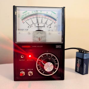 Op de afbeelding: Een zwarte analoge Micronta-multimeter met een helder display dat verschillende schalen toont voor ohm-, gelijkstroom- en wisselstroommetingen. Het apparaat heeft een grote draaiknop, twee tuimelschakelaars, een rood indicatielampje en een 9V-batterij.