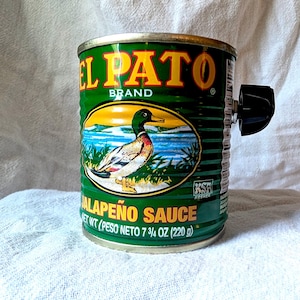 Puede incluir: Una lata de metal verde de salsa de jalapeño marca El Pato con una perilla negra unida al costado. La etiqueta presenta una ilustración de un pato real, el texto EL PATO BRAND, JALAPEÑO SAUCE y NET WT 7 3/4 OZ (220 g) con un símbolo KSA.