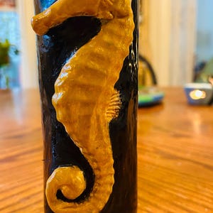 May include: A black ceramic mug with a gold-colored seahorse design. The seahorse has a white eye and a detailed, textured body.