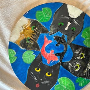 Puede incluir: Un plato de cerámica azul con un diseño pintado a mano de cuatro gatos y un pez dorado. Los gatos son negros, blancos y calicó, y el pez dorado es naranja. El plato está decorado con nenúfares verdes.