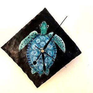 May include: A black square clock with a blue and green painted sea turtle design. The clock hands are black.