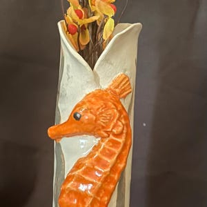 May include: A ceramic vase with a white and gray glaze features a three-dimensional orange seahorse design. The vase is filled with a bouquet of yellow flowers with red berries.