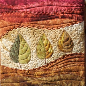 May include: A quilted wall hanging with a landscape design featuring three stylised leaves in shades of green and brown. The background is a gradient of red, orange, brown, and green.