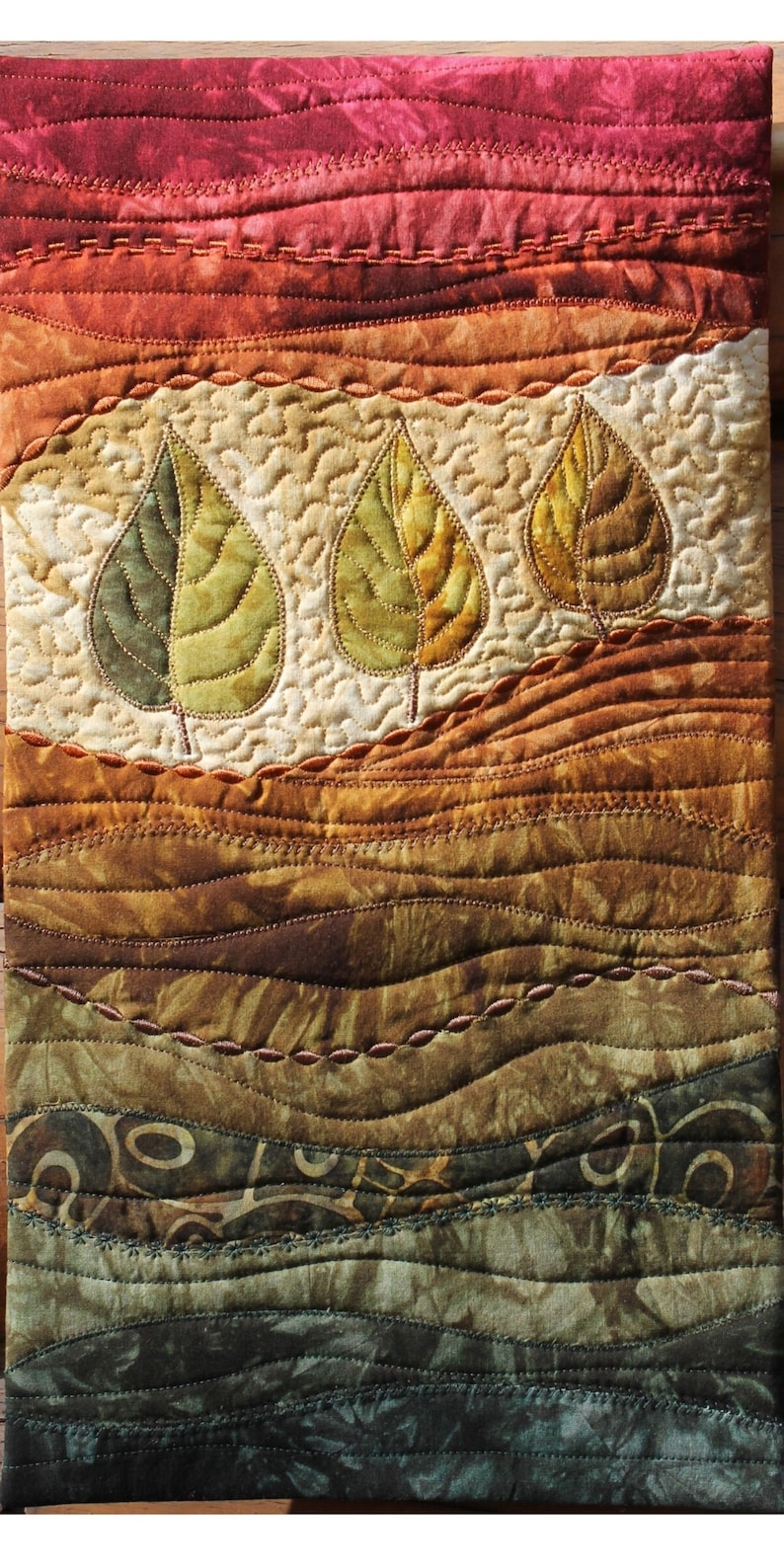 May include: A quilted wall hanging with a landscape design featuring three stylised leaves in shades of green and brown. The background is a gradient of red, orange, brown, and green.
