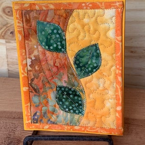 May include: A small quilted wall hanging with a yellow background and three green leaves with white polka dots. The leaves are stitched onto the fabric and the edges are outlined with a silver thread.