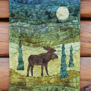 May include: A quilted wall hanging featuring a brown moose standing in a green and yellow landscape with a full moon in the sky.