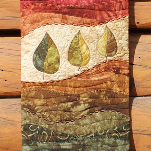 May include: A quilted wall hanging with a landscape design featuring three stylised leaves in shades of green and brown. The background is a gradient of red, orange, brown, and green.
