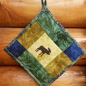 May include: A quilted pot holder with a moose silhouette in the centre. The pot holder is made of green, blue, and yellow fabric with a hanging loop.