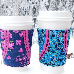 Könnte beinhalten: Zwei weiße Kaffeetassen mit Stoffhüllen. Die Hülle der Tasse links hat ein pinkfarbenes und blaues Blumenmuster. Die Hülle der Tasse rechts hat ein blaues und schwarzes Blumenmuster.