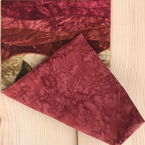 May include: A red and brown quilted fabric placemat with a floral pattern. The placemat is folded in half, with the top half folded over the bottom half.