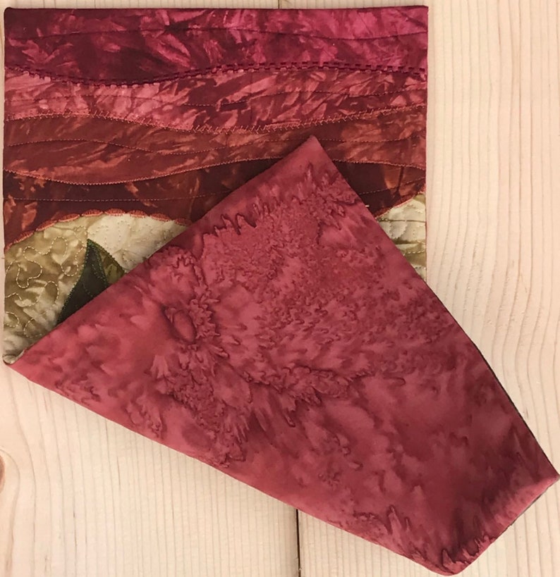 May include: A red and brown quilted fabric placemat with a floral pattern. The placemat is folded in half, with the top half folded over the bottom half.