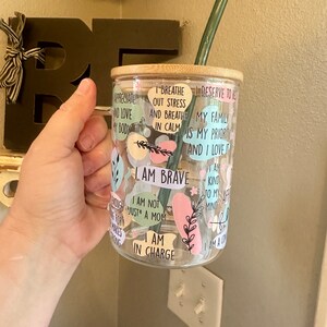 May include: A clear glass tumbler with a bamboo lid and a green reusable straw. The tumbler is decorated with colorful stickers with inspirational quotes, including "I am brave", "I am not just a mom", "I am in charge", "I breathe out stress and breathe in calm", "My family is my priority and I love it", and "I am kind to my mind".