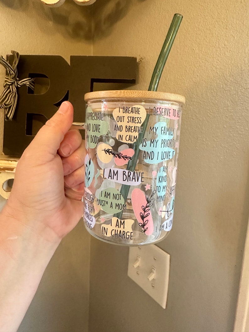 May include: A clear glass tumbler with a bamboo lid and a green reusable straw. The tumbler is decorated with colorful stickers with inspirational quotes, including "I am brave", "I am not just a mom", "I am in charge", "I breathe out stress and breathe in calm", "My family is my priority and I love it", and "I am kind to my mind".
